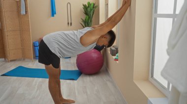 Genç adam sağlık merkezinde yoga minderi, egzersiz topu ve spor ekipmanıyla esniyor.