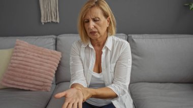 Dekoratif yastıklı rahat bir oturma odasında kanepede otururken bileğindeki ağrıyı değerlendiren bir kadın..
