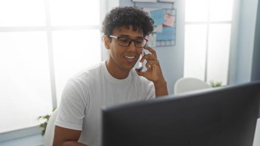 Ofisteki genç adam telefonda konuşurken arka planda evrakları olan bilgisayarda çalışıyor, profesyonel bir iş ortamı sergiliyor.