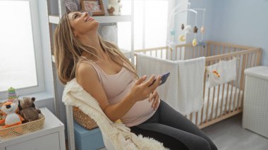 Bebek beşiği ile rahat bir yatak odasında telefonda konuşan hamile bir kadın, huzurlu ve samimi bir ev atmosferini yansıtıyor..