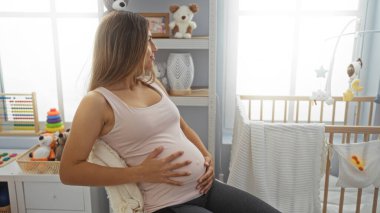 Bebek beşiği ve oyuncaklarıyla rahat bir kreşte oturan hamile bir kadın, bebek beşiğine bakıyor, evinin huzurlu atmosferinin tadını çıkarıyor, ve içeriden memnuniyet duyuyor..