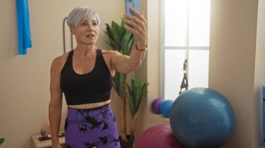 Orta yaşlı, kısa gri saçlı bir kadın spor salonunda selfie çekiyor. Etrafında spor aletleri var.