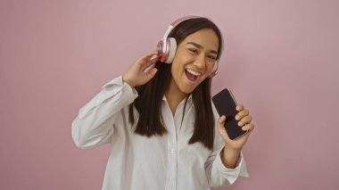 Pembe kulaklıklı müzik dinleyen genç bir kadın ve izole edilmiş pembe bir arka plan üzerinde akıllı telefon tutan bir kadın.