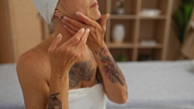 Boynuna krem süren bir kadın, sakin bir spa ortamında, kollarında dövmeler olan, beyaz bir havlu takmış, güzellik ve sağlığını ön plana çıkaran bir kadın.