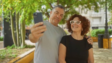 Şehir parkında birlikte selfie çeken orta yaşlı bir çift, açık havada sevgi ve mutluluk sergiliyorlar, canlı, kentsel bir geçmişleri var..