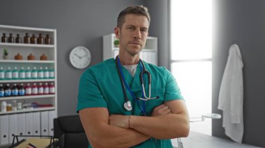 Hastane odasında steteskobu olan kendinden emin bir erkek doktor gün boyunca tıbbi malzeme rafı ve duvar saatinin önünde kolları çapraz duruyor.