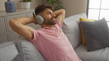 Sakallı genç bir adam evinde kanepede dinleniyor, rahat bir oturma odasında kulaklıkla müzik dinliyor..