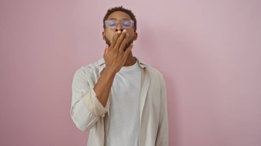 Gözlüklü genç adam ağzı kapalı esniyor, pembe bir arka plana karşı duruyor, günlük kıyafetler giyiyor..