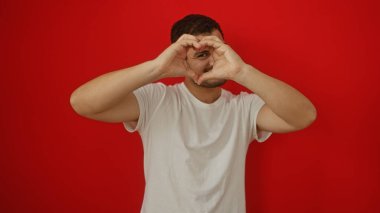 Yakışıklı, genç, İspanyol bir adam elleri ile kalp şekli oluşturan izole edilmiş kırmızı arka plan sevgi ve sevgi gösteriyor.