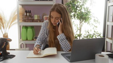 Dizüstü bilgisayarlı ve arka planda ürünleri olan bir mağazada telefonla konuşurken deftere yazan genç bir kadın.