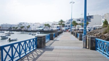 Ispanya 'nın kanarya adalarında sakin bir açık hava manzarası yakalayan, net bir gökyüzü altındaki odak binaları ve teknelerden oluşan, mavi bir korkuluk ve limanlı Lanzarote' deki bir sahil yürüyüşünün bulanık görüntüsü.