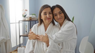 Beyaz elbiseli bir anne ve kız güzellik merkezine gülümsüyor ve sarılıyorlar, kapalı bir sağlık ortamında sevgi dolu bir aile anını yakalıyorlar.
