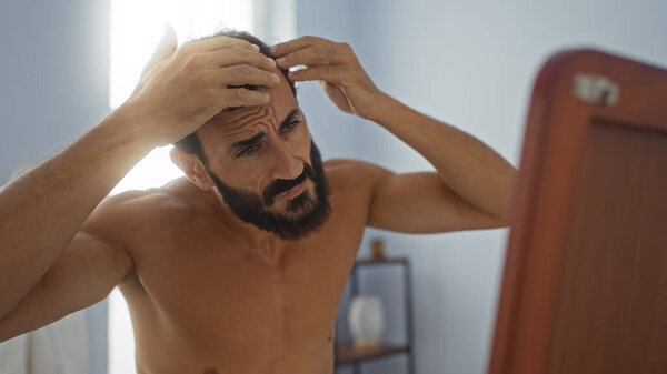 Молодой человек осматривает волосы в зеркале в закрытом оздоровительном центре, выглядя обеспокоенным в салоне красоты.