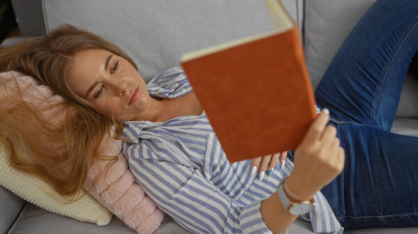 Молодая женщина читает книгу, расслабляясь на диване в своей уютной гостиной, в полосатой рубашке и джинсах.