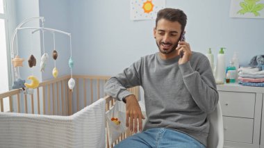 Genç adam rahat bir bebek odasında oturuyor, telefonda konuşuyor, etrafı beşik ve oyuncaklarla çevrili, evde huzurlu ve modern bir aile ortamı yaratıyor..
