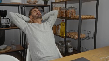 Young man relaxing in office workspace with hands behind head surrounded by shelves of food items and kitchenware, conveying a casual and serene atmosphere indoors. clipart