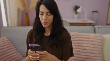 Rahat bir oturma odasında akıllı telefon kullanan genç bir kadın, şık dekorları, yastıkları ve ev yapımı iç dekorasyonları olan çekici bir İspanyol kadını içeriyor..