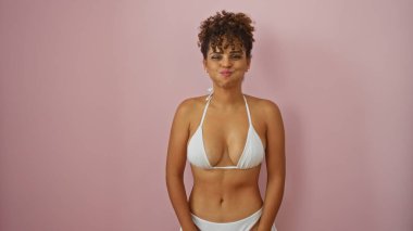 Beyaz bikinili genç bir kadın izole edilmiş pembe bir arka plana karşı poz veriyor, esmer kıvırcık saçlarıyla güzel ve kendinden emin bir yüz ifadesi sergiliyor.