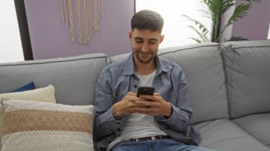 Yakışıklı İspanyol bir adam rahat bir kanepede rahat bir şekilde dinleniyor modern oturma odasında gülümseyen bir akıllı telefon kullanıyor boş zamanlarını ve bağlantılarını gösteriyor..