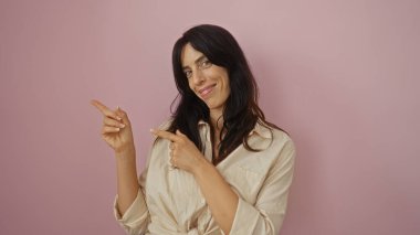 Genç bir kadın gülümsüyor ve pembe bir arka planı işaret ediyor.