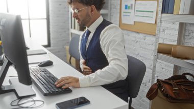 Ofisteki genç adam iş belgeleriyle çevrili bilgisayarla masa başında otururken midesi bulanıyor. Bu da iş baskısı ve sağlık sorunları olduğunu gösteriyor.