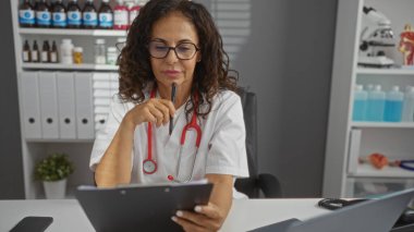 Mature female doctor with glasses in a hospital office, thoughtfully holding a pen while examining documents, surrounded by medical supplies and equipment. clipart