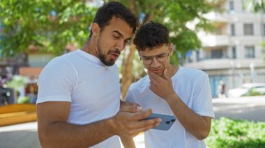 Baba ve oğul birlikte telefona bakıyorlar. Şehir parkında şaşırmış bir ifade var.