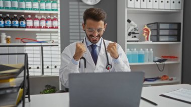 Gözlüklü genç adam klinikte kutlama yapıyor, laboratuvar önlüğü giyiyor, tıbbi malzemeler ve bilgisayarla çevrili, evde olumlu duygular ve profesyonellik sergiliyor..