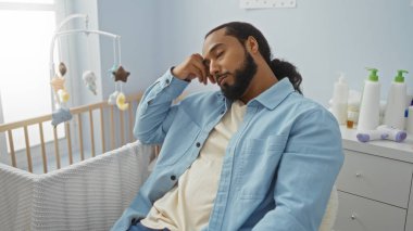Sakallı genç adam bebek odasında uyuyor, günlük giysiler giyiyor, bebek eşyalarıyla çevrili, gündüzleri huzurlu bir ev ortamında, yorgunluğu yansıtıyor..