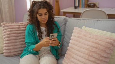Oturma odasında oturan genç bir kadın rahat bir koltukta akıllı telefon kullanıyor. Arka planda yastıklar ve masa var. Kapalı, çekici ve odaklı..
