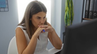 Ofis odasında bilgisayarlı ve arkaplanda dosyaları olan İspanyol bir kadın odaklanmış görünüyor.