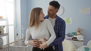 Bebek beşiği ve oyuncaklarıyla çevrili sıcak yatak odasında sevgi dolu kucaklaşan çift, ev ortamında bir erkek ve kadınla sıcak bir aile ortamı olduğunu gösteriyor..