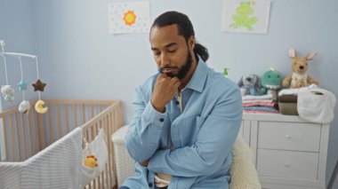 Yatak odasında sakallı genç bir adam beşiğin yanında oturuyordu. Huzurlu, hafifçe aydınlatılmış iç mekânda düşünceli görünüyordu..