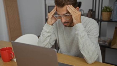 Ofis ortamında dizüstü bilgisayarı ve kırmızı kahve fincanı olan İspanyol bir adam, kafasını iki eliyle tutarken, gözlük ve süveter giyerken derin düşüncelere dalmıştı..