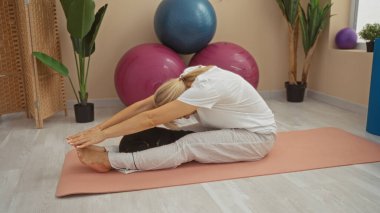 Orta yaşlı sarışın kadın içeride yoga minderinin üzerinde jimnastik salonunda jimnastik topları ve arka planında bitkiler olan bir chihuahua ile geriniyor.