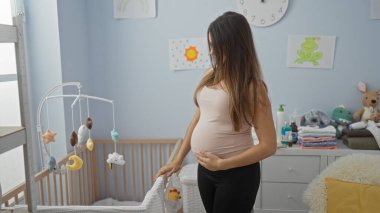 Yatak odasında hamile bir kadın bebek oyuncaklarıyla beşiğe bakıyor. Etrafı güzel ışıklandırılmış ev dekorasyonu ile çevrili. Huzurlu bir annelik anı yakalıyor..