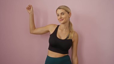 İzole edilmiş pembe bir arka plana karşı güvenle poz veren bir kadın atletik yapısını ve sarı saçlarını dostça ve çekici bir tavırla sergiliyor..