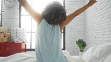 Beyaz tuğlalı duvarları ve geniş pencereleri olan, rahat bir yatak odasında kıvırcık saçlarıyla yatakta uzanan bir kadın.