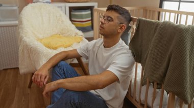 Genç adam düşünceli bir şekilde bebek beşiği ve yumuşak mobilyalarla çevrili rahat bir bakımevinde oturuyor, günlük giysiler ve gözlük takıyor, evde sakin bir tavır sergiliyor..
