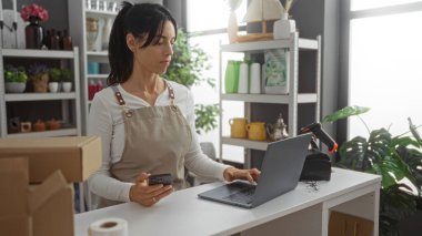 Dükkanda çalışan, etrafı dekorasyon elemanlarıyla çevrili, dizüstü bilgisayar kullanan, önlük giyen, İspanyol, yetişkin, paketleri açan, rafları, kartonları, iç mekanı olan bir kadın.