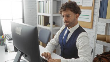 Resmi kıyafetler giyen genç bir adam, kitap ve belgelerle çevrili modern bir ofis ortamında bilgisayarda çalışıyor, odaklanmış bir iş ortamını gösteriyor..