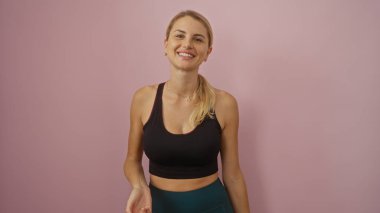 İzole edilmiş pembe duvara gülümseyen, güzellik ve mutluluğu resmeden, spor kıyafetleri ve sarı saç stiliyle gündelik moda sergileyen, kadınlığı vurgulayan bir kadın..