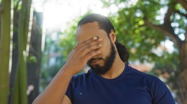 Sakallı genç adam şehir parkı ortamında endişeli görünüyor, yüzünü güneş ışığı altında eliyle kapatıyor, bir anlık stres ya da düşüncelere dalma anı yakalıyor.