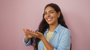 Güzel, genç, İspanyol bir kadın, mavi gömlekli ve parlak bir şekilde gülümseyen, izole edilmiş pembe bir arka plan duvarına el kol hareketi yaparak para harcıyor.