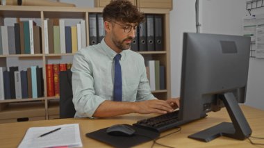 Young man working on computer in office with bookshelves behind, focused on typing, wearing glasses and tie, indoor setting indicating workplace or professional environment clipart