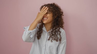 Genç bir kadın bir eliyle yüzünü kapatarak gülüyor, pembe bir arka planda duruyor, çizgili bir gömlek giyiyor, mutluluğu ve oyunculuğu iletiyor.