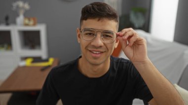 Genç İspanyol bir adam rahat bir oturma odasında gözlük ayarlıyor, rahat ve şık bir iç mekan atmosferi sergiliyor..