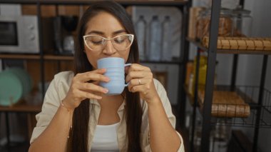 Kadın, rahat bir oturma odası ortamında kahveden zevk alıyor. Rahat ve rahat bir atmosfer yaratıyor. Arka planda şık bir dekorasyon var..