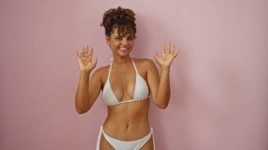 İzole edilmiş pembe bir arka plan üzerinde bikinili bir kadın, güzel kıvırcık saçlarını ve kendine güvenen gülümsemesini gösteriyor, Latin görünümlü genç, yetişkin, İspanyol bir kadını yansıtıyor..