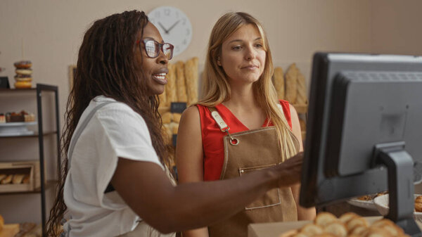 Two women, bakery workers in aprons, stand indoors in front of a computer monitor in a bakery shop, surrounded by bread and bakery products.